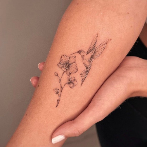 A fine-line tattoo of a hummingbird in flight near a blooming flower on a person's inner forearm, showcasing intricate black-and-gray detailing.